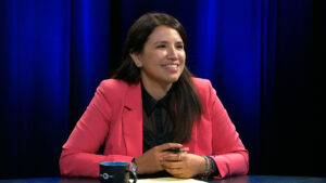 A woman in a pink blazer sits at a table with a notepad and a mug, smiling, against a blue curtain background.