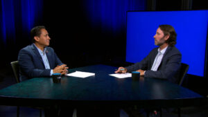 Two men in suits sit across from each other at a round table with papers and cups, engaged in conversation in a studio setting with a blue backdrop.