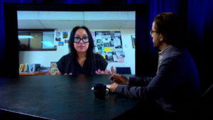 A man sits at a round table with a coffee mug, speaking via video call to a woman displayed on a large screen in a studio setting.