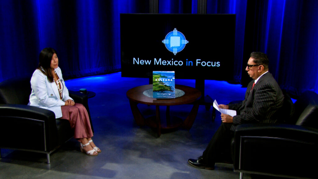 Two people sit in armchairs having a conversation in a TV studio with a "New Mexico in Focus" sign displayed on a screen behind them.