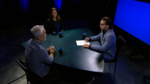 Three people sit around a table under studio lights, with one person holding papers and appearing to lead the discussion. Three mugs and a notepad are on the table.