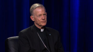 An older man in religious attire, including a clerical collar and a cross necklace, sits in a chair against a dark blue background.