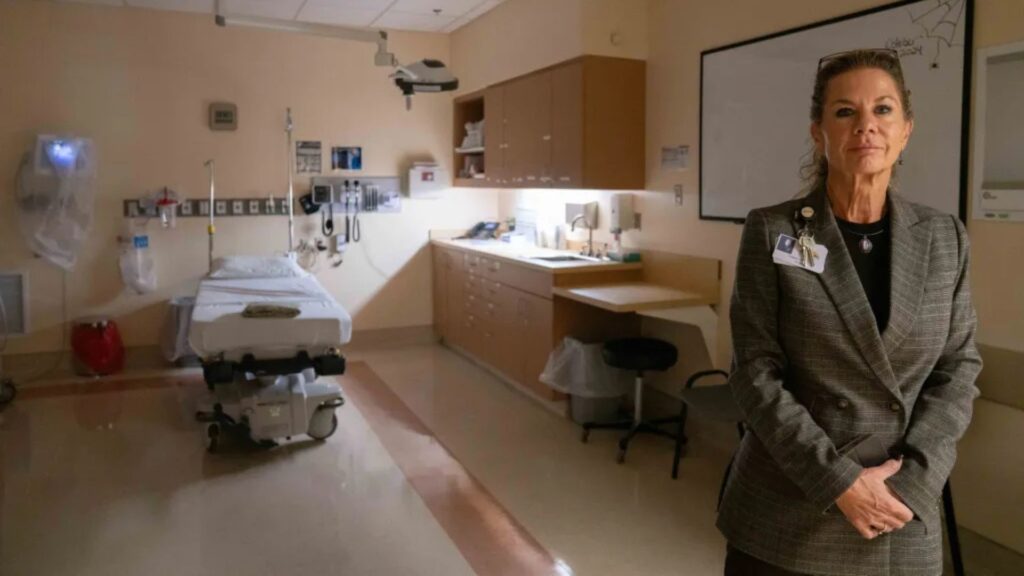 A woman in a suit stands in a hospital room next to an empty bed and medical equipment.