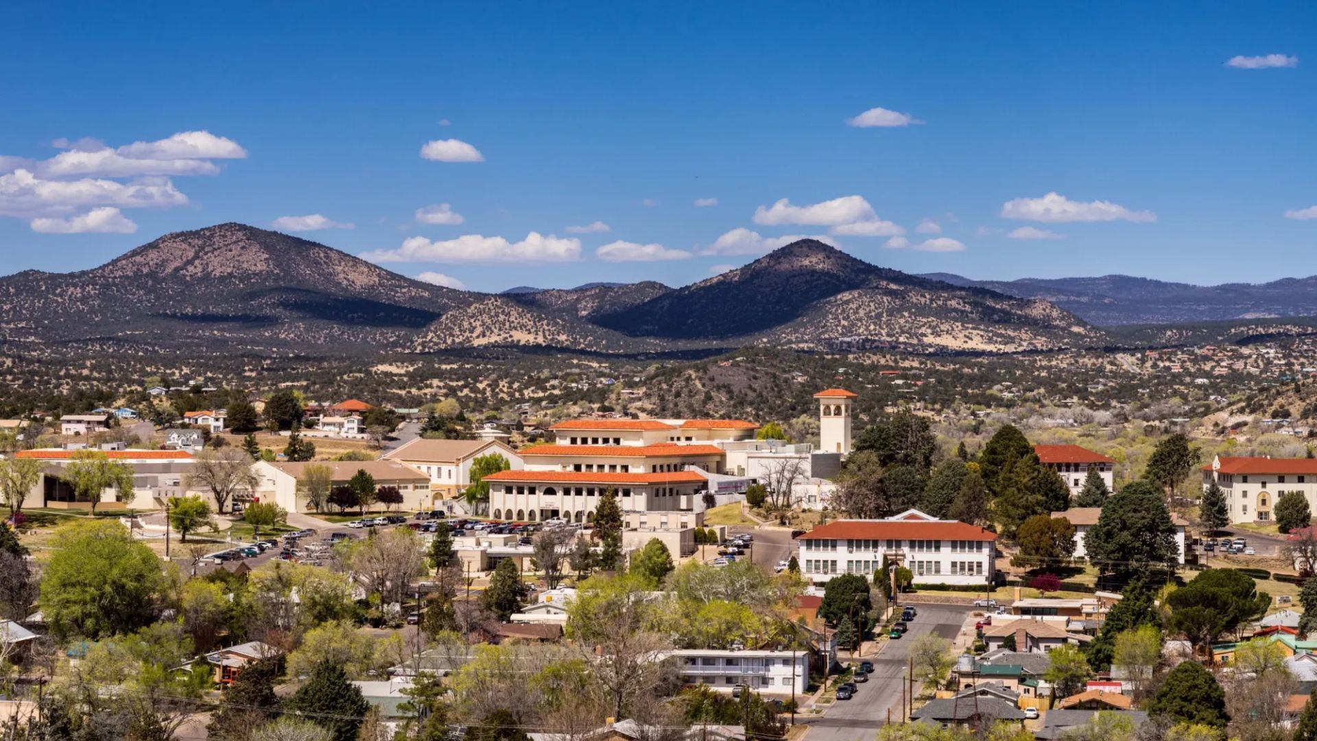 A small town with red-roofed buildings and tree-lined streets set against forested hills and mountains under a clear blue sky.