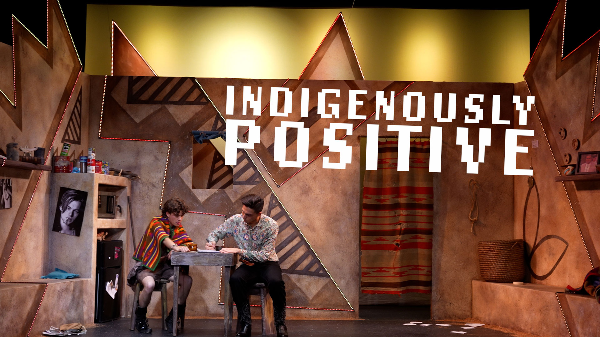 Two people sit at a small table on a stage set with geometric patterns, shelves, and photos. Large text reads "Indigenously Positive" above them.