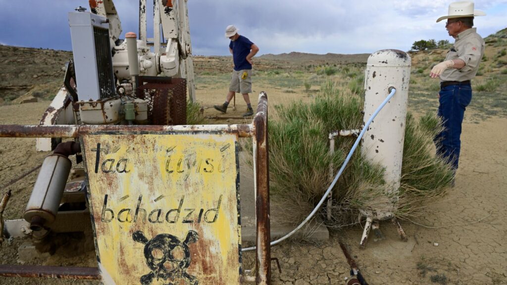 Two men stand near old, rusted oil equipment in a dry, barren landscape; a warning sign with a skull symbol and text is visible in the foreground.