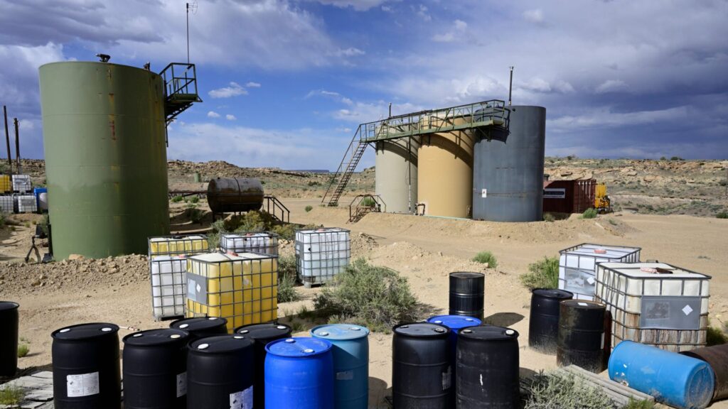 Industrial storage tanks and various plastic and metal barrels are arranged on a dirt lot in a semi-arid landscape under a partly cloudy sky.