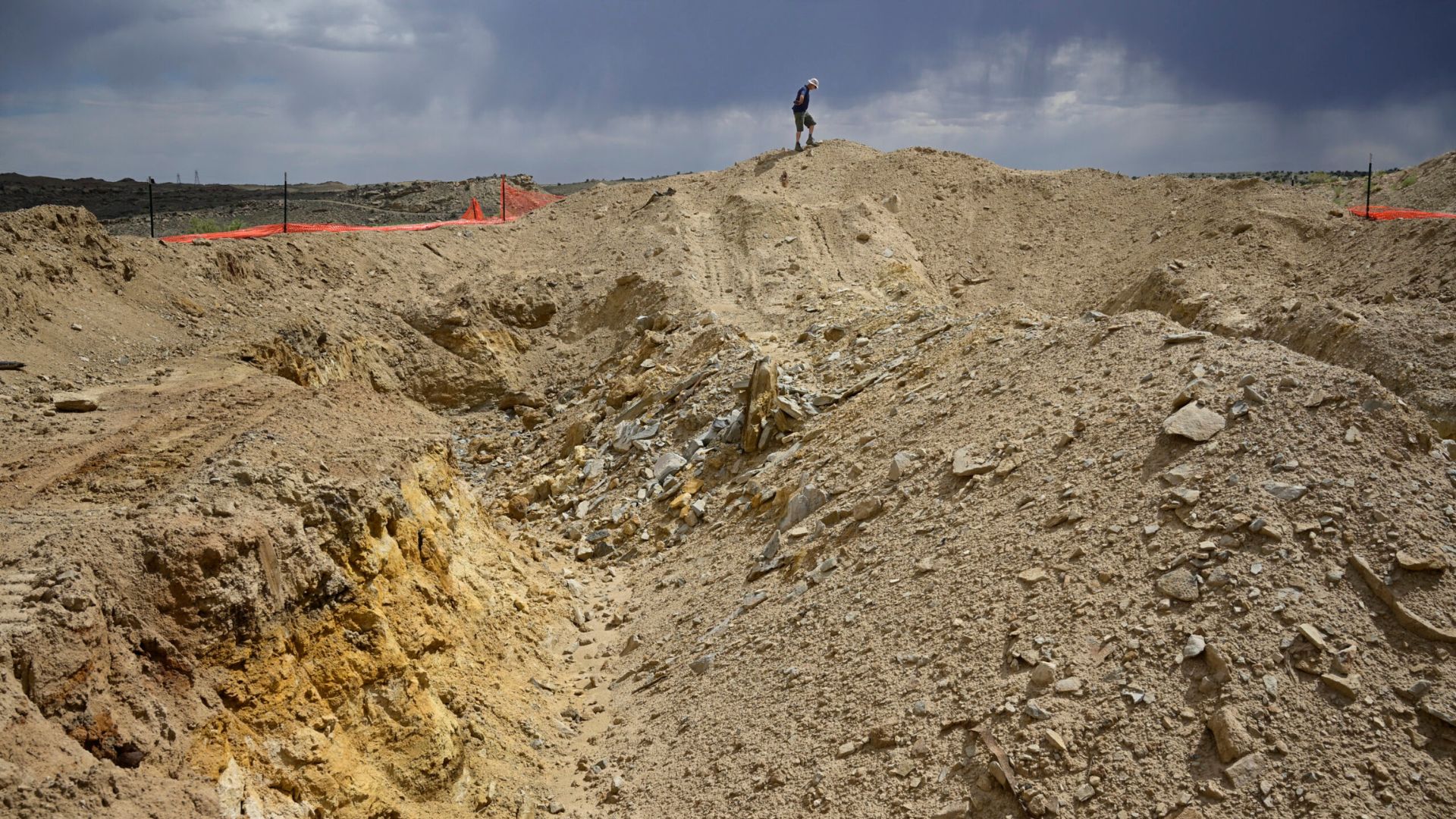 A person stands atop a large mound of earth, surrounded by trenches and rocky soil under a cloudy sky; orange safety netting is visible in the background.