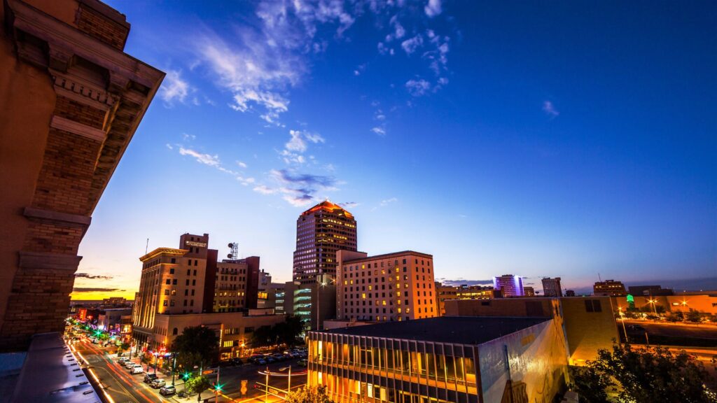 Cityscape at dusk showing illuminated streets and mid-rise buildings under a blue sky with scattered clouds.