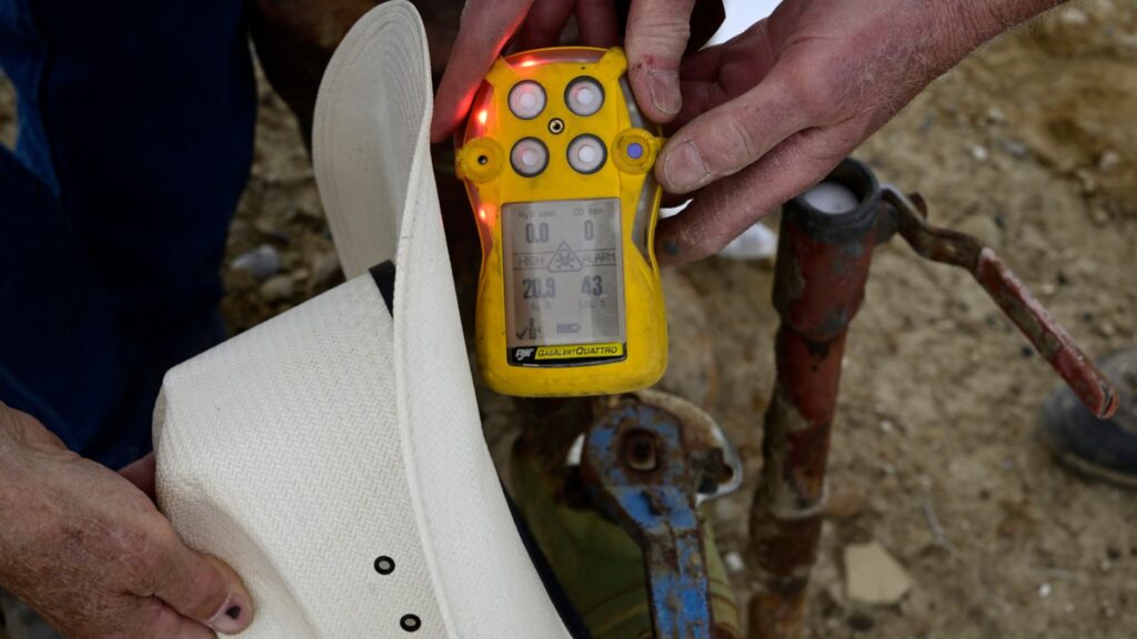 A person holds a yellow radiation detector near a white hat and a metal object outdoors, showing measurement readings on the device’s screen.