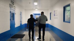 Two men walk down a brightly lit hallway labeled “Food Service”; two more people stand at the end of the hall near a door.