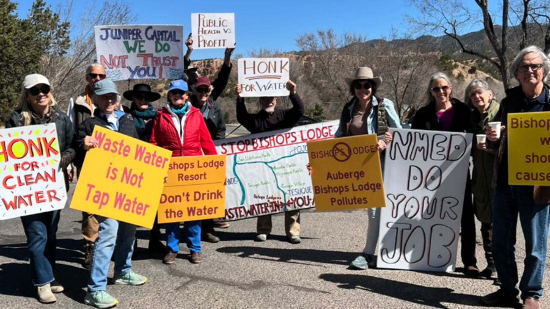 A group of people stand outdoors holding signs protesting for clean water and against wastewater, including messages like “Honk for Clean Water” and “NMED Do Your Job.”.