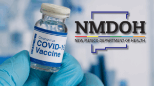 A gloved hand holds a vial labeled "COVID-19 Vaccine" next to the New Mexico Department of Health (NMDOH) logo.
