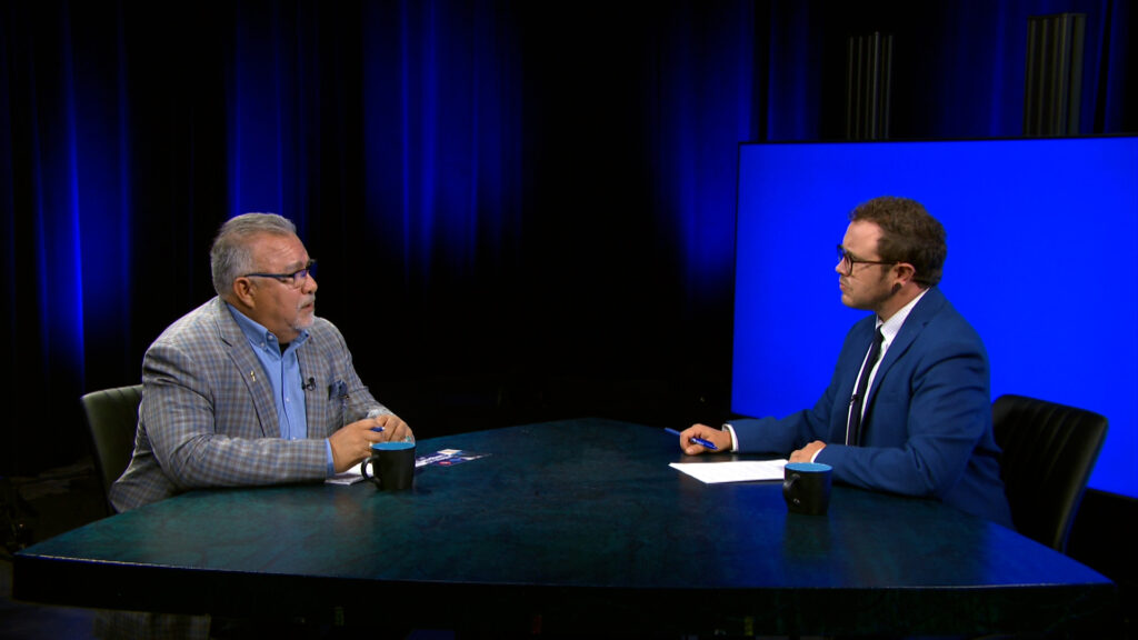 Two men in suits sit across from each other at a table in a studio with blue lighting, engaged in conversation with papers and coffee mugs on the table.