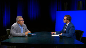 Two men in suits sit across from each other at a table in a studio with blue lighting, engaged in conversation with papers and coffee mugs on the table.