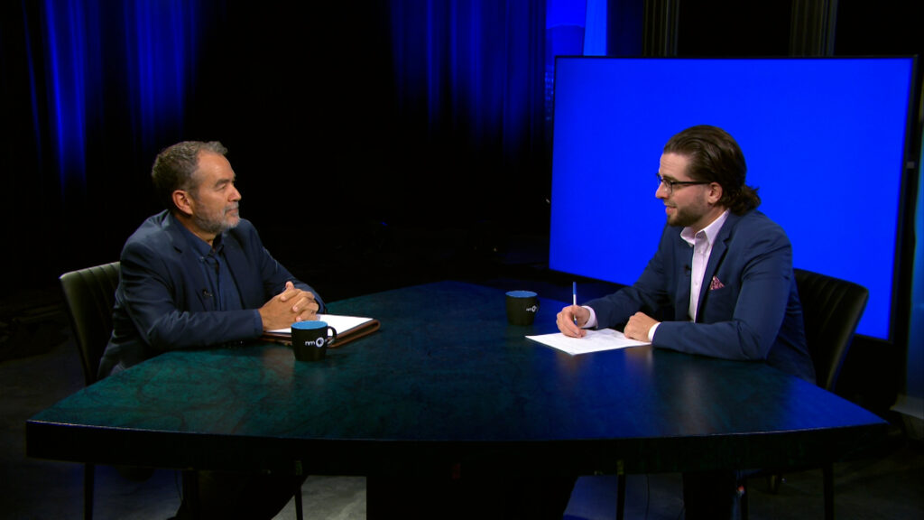 Two men in suits sit across from each other at a table in a studio, one holding a notebook and the other writing on paper, with a blue screen in the background.