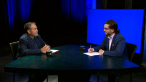 Two men in suits sit across from each other at a table in a studio, one holding a notebook and the other writing on paper, with a blue screen in the background.