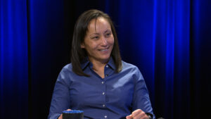 A person with straight dark hair and a blue button-up shirt sits at a table, smiling, against a background with dark blue curtains.
