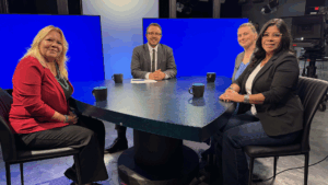 Four people sit around a round table with black coffee mugs in a studio setting with bright blue screens in the background.