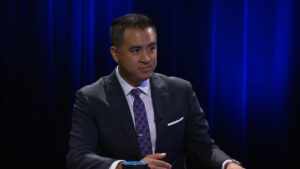 A man in a suit and tie sits at a table against a dark blue curtain backdrop, holding a pen and a blue cup is visible in front of him.