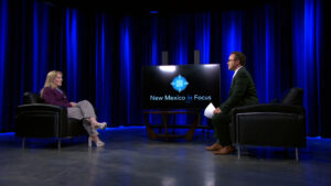 A woman and a man sit facing each other in a studio with blue curtains, with a screen between them displaying "New Mexico in Focus.