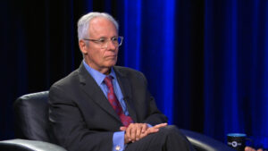 An older man in a suit sits in a black armchair on a studio set with blue lighting, looking ahead with a serious expression.