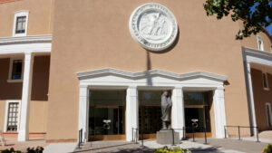 The exterior of a government building with the New Mexico state seal above the entrance and a statue in front.
