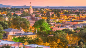 Aerial view of downtown Santa Fe, New Mexico at sunset, featuring adobe buildings, lush trees, and a prominent tower with a flag.