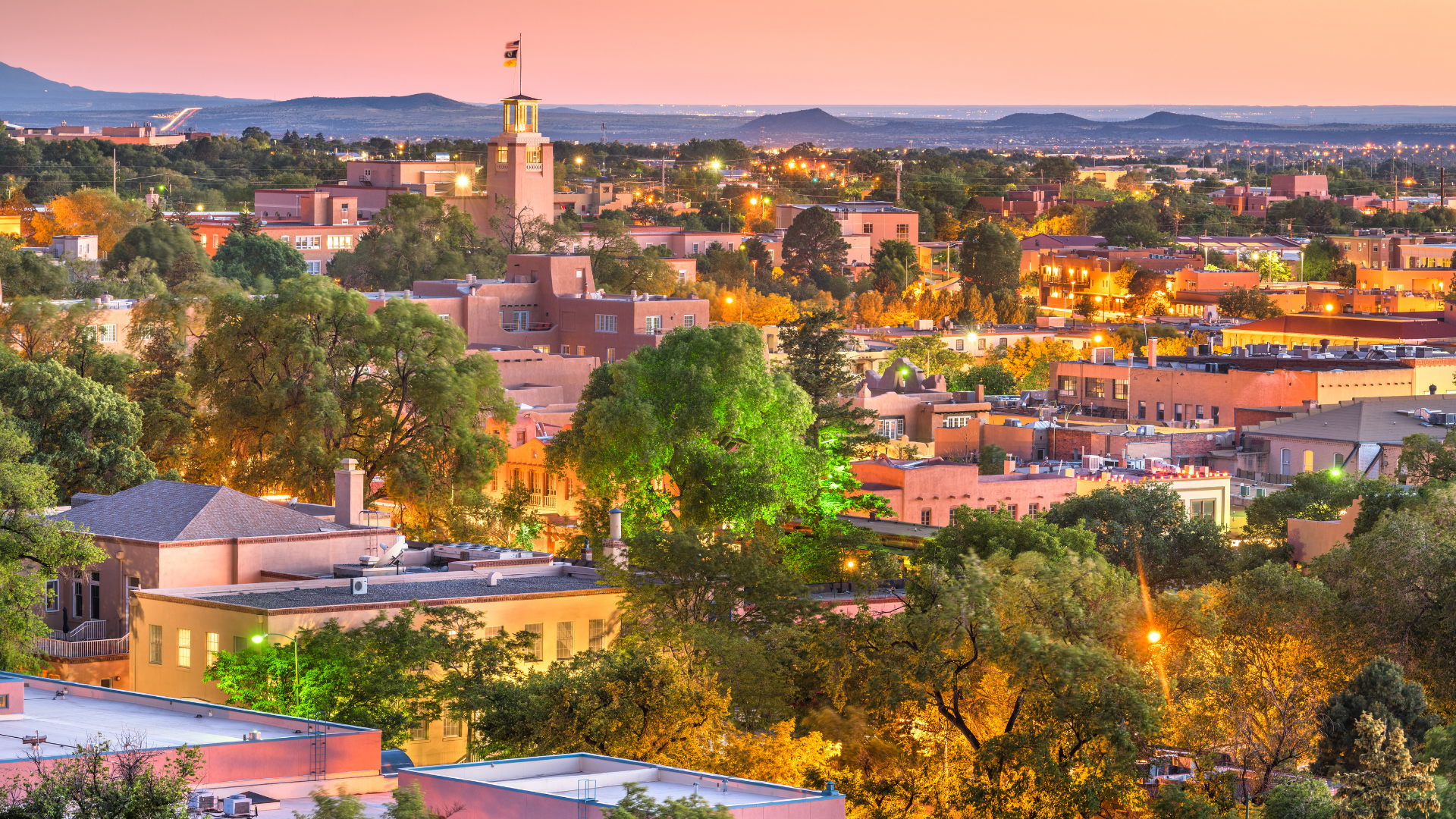 Aerial view of downtown Santa Fe, New Mexico at sunset, featuring adobe buildings, lush trees, and a prominent tower with a flag.