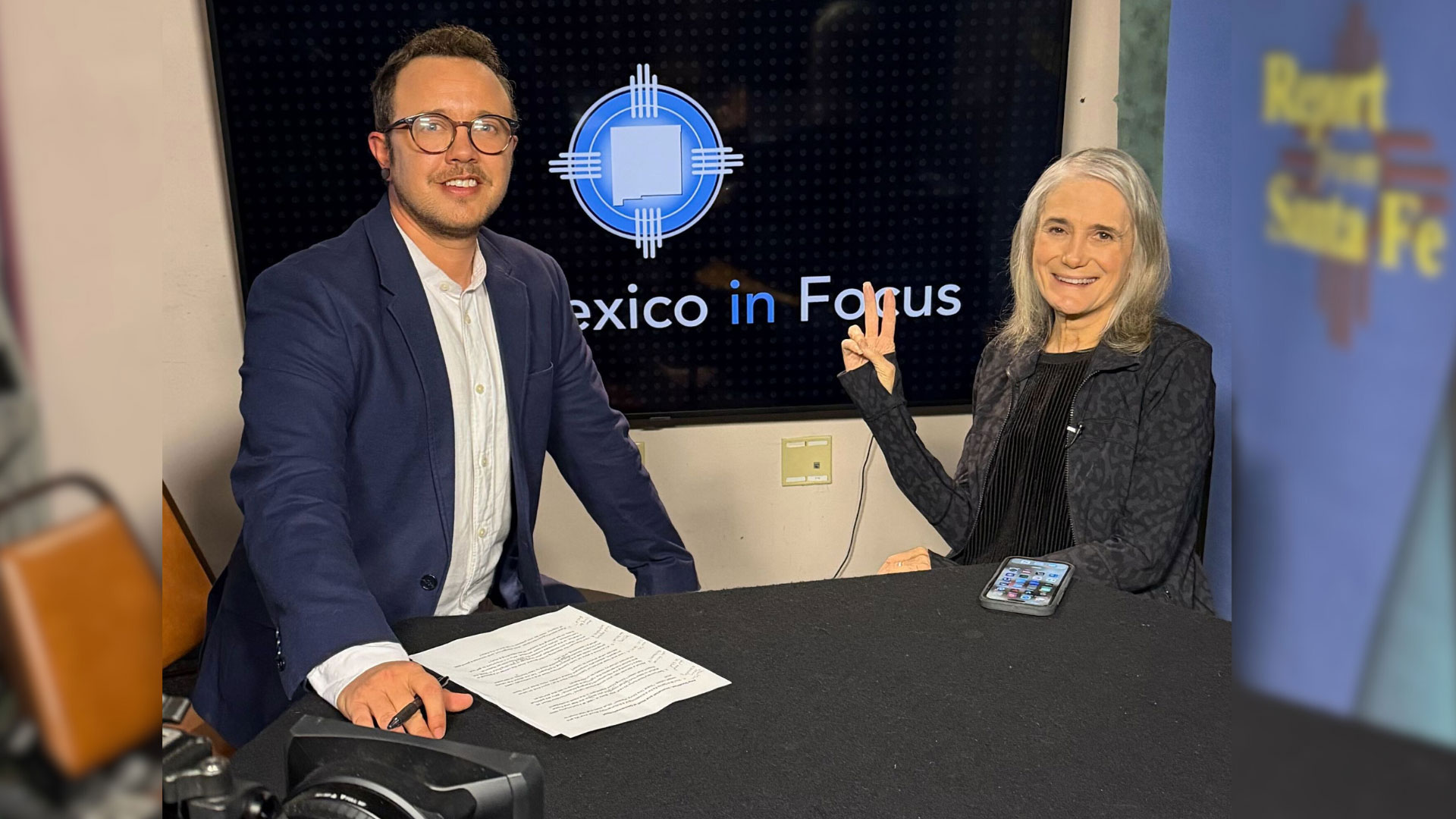 Two people sit at a table in a studio setting with "Mexico in Focus" displayed on a screen behind them; one has papers, the other is smiling and holding up two fingers.