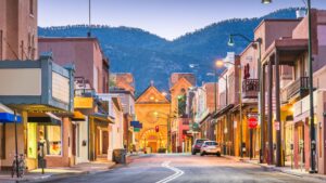A quiet street in a small town with adobe-style buildings, parked cars, and a historic church in the background, set against a backdrop of tree-covered hills.