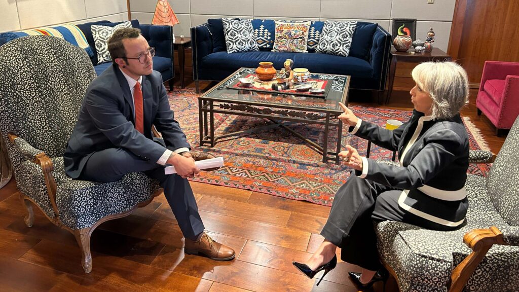 Two people sit in armchairs, engaged in a serious conversation in a formal living room setting with a coffee table and patterned decor.
