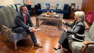 Two people sit in armchairs, engaged in a serious conversation in a formal living room setting with a coffee table and patterned decor.