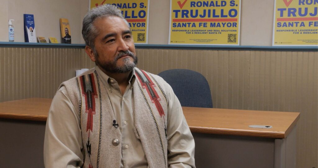 A man with gray hair and beard sits in an office with Ronald S. Trujillo for Santa Fe Mayor campaign posters on the wall behind him.