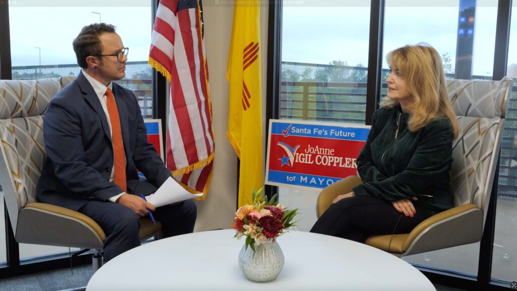A man in a suit and a woman in a dark green jacket sit across from each other in an office. Campaign signs and flags are visible in the background.