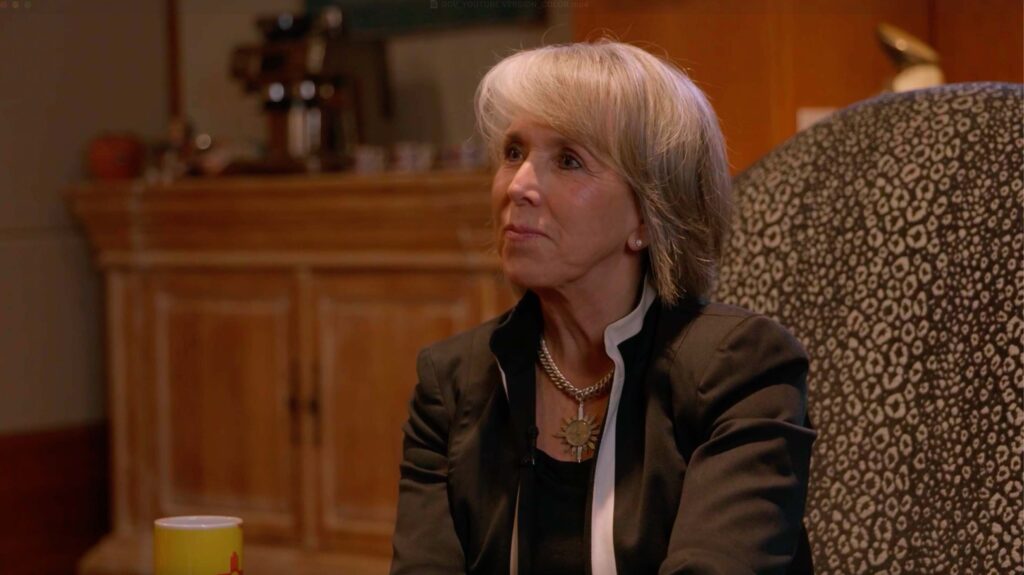 A woman with short gray hair and a black blazer sits indoors near a patterned chair, with a wooden cabinet and a cup in the background.