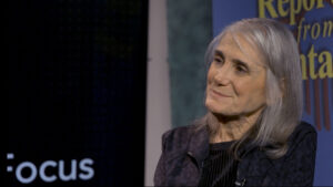 An older woman with long gray hair sits in front of a colorful background, looking slightly to the side and smiling gently. The words "focus" and "Report from Santa" are partially visible.