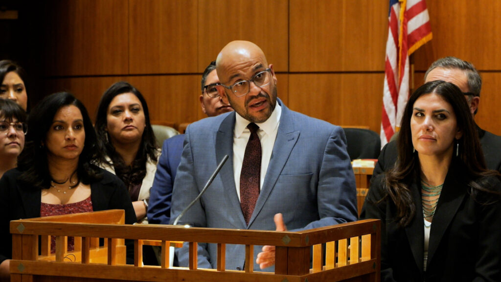 A man in a blue suit speaks at a podium in a courtroom, surrounded by several people, with an American flag in the background.