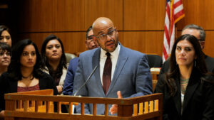 A man in a blue suit speaks at a podium in a courtroom, surrounded by several people, with an American flag in the background.