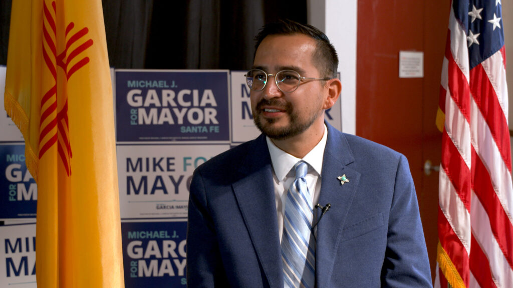 A man in a blue suit and tie sits indoors with campaign signs reading "Garcia for Mayor" in the background, near American and New Mexico flags.