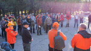 A group of people, many in orange hats and jackets, gather outdoors for a briefing or meeting in a wooded area.