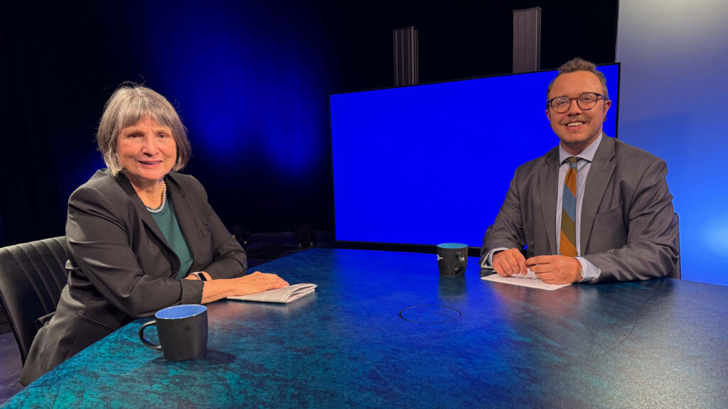 Two people sit at a round table with notebooks and mugs, facing the camera. A large blue screen is in the background in a studio setting.