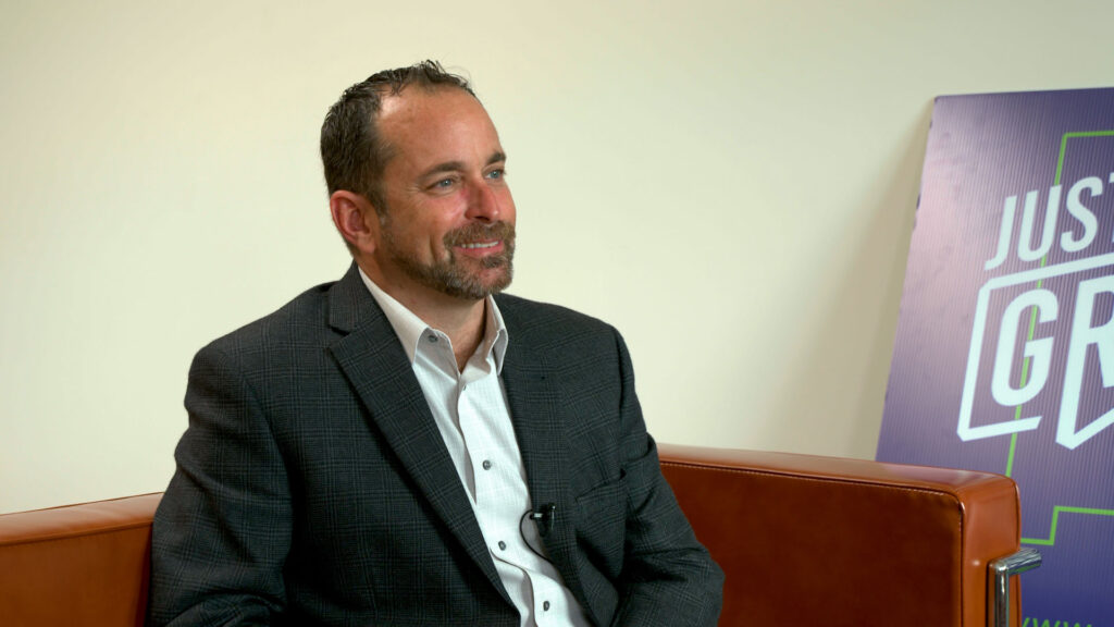 A man in a dark blazer and white shirt sits on a brown leather chair, smiling, with a purple and white sign in the background.