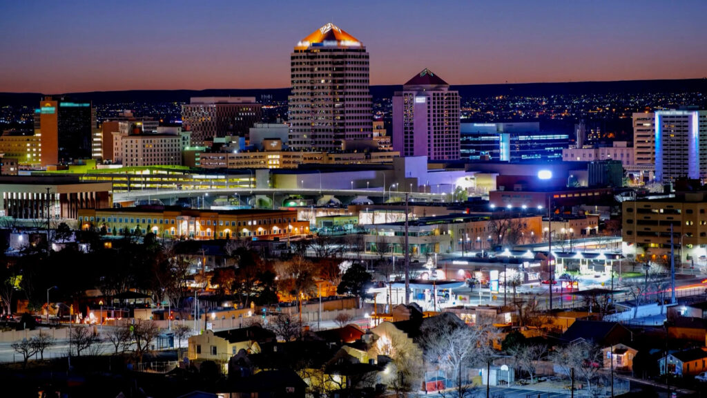 Cityscape at dusk showing downtown Albuquerque, New Mexico, with illuminated buildings and streets against a darkening sky.
