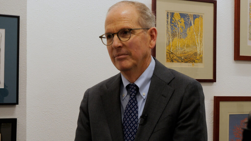 A man wearing glasses and a dark suit sits in front of a white wall with framed artwork.