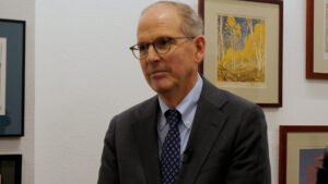 A man wearing glasses and a dark suit sits in front of a white wall with framed artwork.