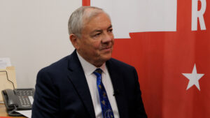 Older man in a dark suit and patterned blue tie sits indoors near a red and white backdrop, with a phone and papers visible in the background.