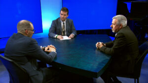 Three men in suits sit around a round table having a discussion in a studio with blue screens in the background.