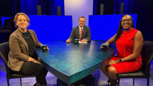 Three people sit at a triangular table in a studio with blue screens behind them, each holding a mug and facing the camera.