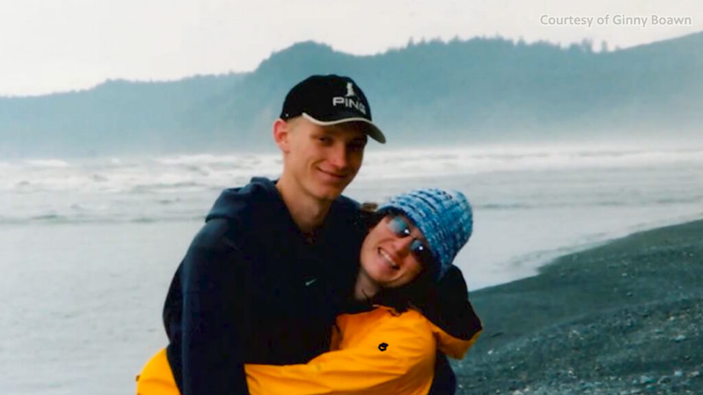 Two people stand on a rocky beach; one wears a black cap and dark hoodie, while the other, in a blue hat and yellow jacket, hugs them with misty hills and ocean in the background.